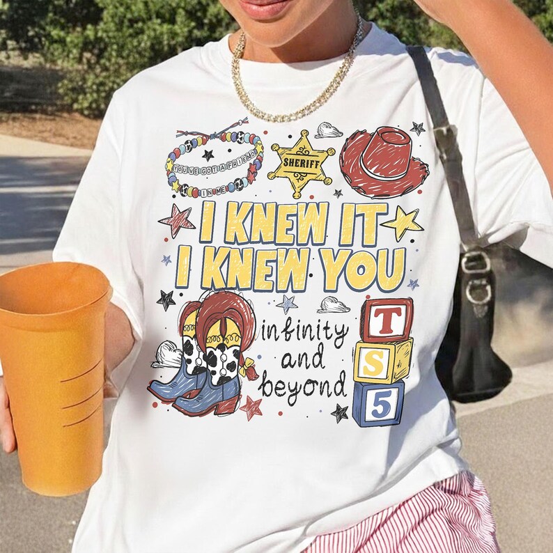 May include: A white t-shirt featuring a colorful graphic print with the text I KNEW IT I KNEW YOU and infinity and beyond. The design includes illustrations of a beaded bracelet, a yellow sheriff star, a red cowboy hat, cowboy boots, and blocks.