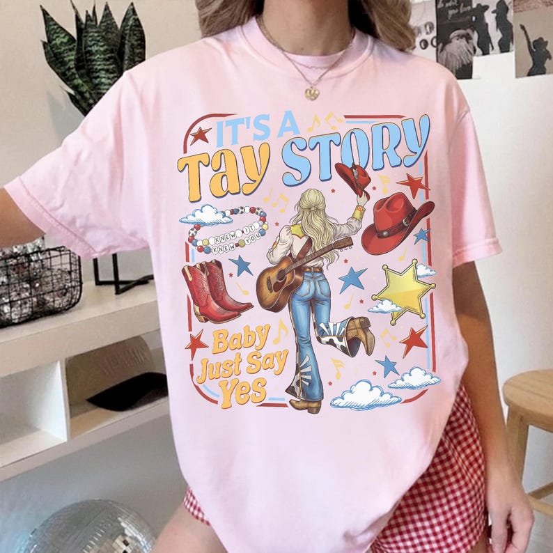 May include: A light pink t-shirt featuring a graphic print with the text IT'S A TAY STORY and Baby Just Say Yes. The design includes a person playing a guitar, red cowboy boots, a red hat, a beaded bracelet, stars, and clouds in a square frame.