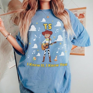 May include: A light blue t-shirt featuring a graphic of a cowgirl character playing an acoustic guitar. The design includes white clouds, musical notes, a ball, a traffic cone, and the yellow text TS and I Knew It, I Knew You.