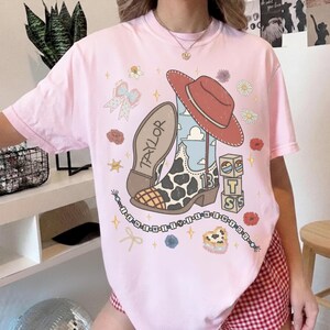 May include: A light pink t-shirt featuring a graphic print of a cowboy boot with a cow print pattern, a red cowboy hat, alphabet blocks, and a beaded string with the text I KNEW IT I KNEW YOU. The boot has the name TAYLOR and number 13.