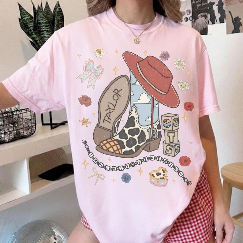 May include: A light pink t-shirt featuring a graphic print of a cowboy boot with a cow print pattern, a red cowboy hat, alphabet blocks, and a beaded string with the text I KNEW IT I KNEW YOU. The boot has the name TAYLOR and number 13.