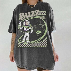 May include: Dark gray t-shirt featuring a graphic of Buzz Lightyear from Toy Story. The design includes the word "Buzz" in a retro font, the year "1995", and a checkered pattern. The shirt has short sleeves and a relaxed fit.