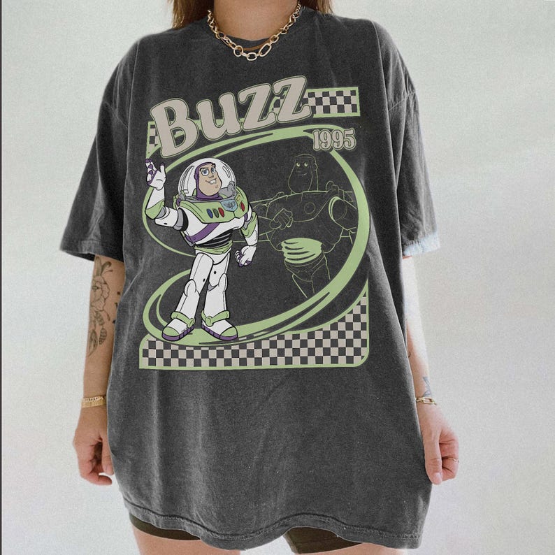 May include: Dark gray t-shirt featuring a graphic of Buzz Lightyear from Toy Story. The design includes the word "Buzz" in a retro font, the year "1995", and a checkered pattern. The shirt has short sleeves and a relaxed fit.