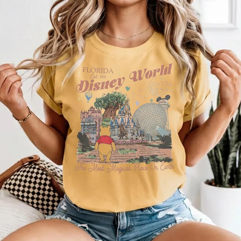 Puede incluir: Camiseta de color amarillo mostaza con un gr&aacute;fico de Disney World en Florida. El dise&ntilde;o incluye ilustraciones de Winnie the Pooh, el &Aacute;rbol de la Vida y Spaceship Earth. El texto en la camiseta dice "Florida Disney World" y "The Most Magical Place On Earth."