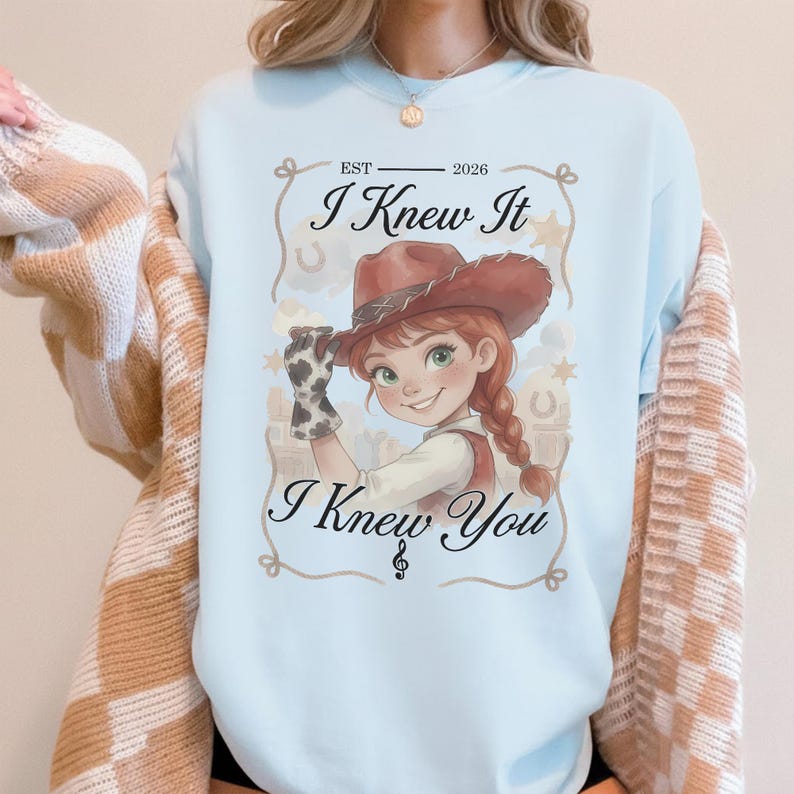 May include: A light blue sweatshirt featuring a graphic of a cartoon cowgirl in a brown hat and cow-print glove. The text reads EST 2026, I Knew It, and I Knew You, with a musical note symbol. The sweatshirt is worn under a tan and white checkered cardigan.
