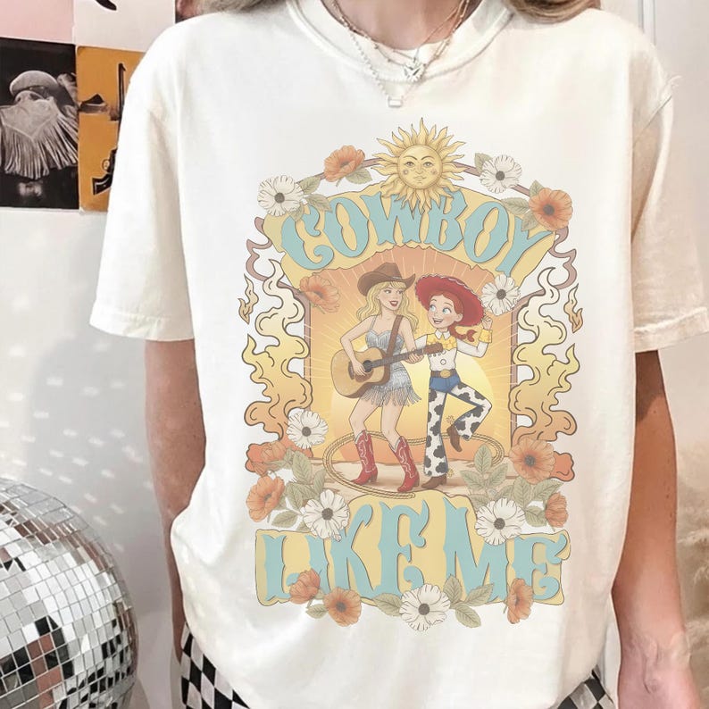 May include: A cream-coloured short-sleeve t-shirt featuring a graphic print of a blonde person playing an acoustic guitar and a red-haired person in a cowboy hat. The text reads COWBOY LIKE ME, surrounded by a sun, flowers, and wavy flame-like borders.