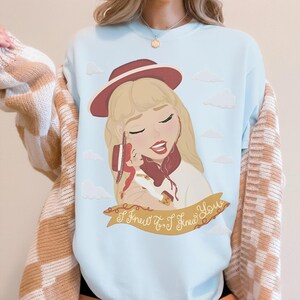 May include: A light blue crewneck sweatshirt featuring a graphic illustration of a blonde character wearing a red hat, holding a smaller doll, with white clouds in the background and a tan banner containing the text I Knew It, I Knew You.