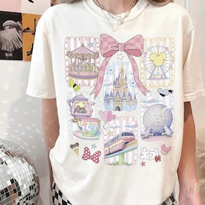 May include: Cream-colored t-shirt featuring a colorful collage of theme park illustrations. The design includes a carousel, castle, Ferris wheel, teapot, teacup, and other whimsical elements. A large pink bow is centered at the top of the design.