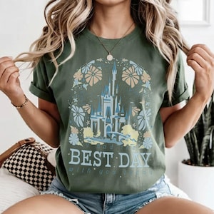 May include: Olive green t-shirt featuring a castle design with blue and yellow accents, surrounded by floral elements. The text "had the BEST DAY with you today" is printed below the castle. The shirt has rolled-up sleeves.