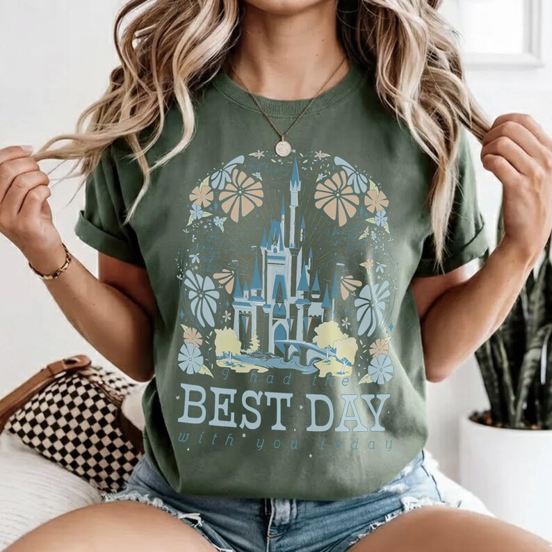May include: Olive green t-shirt featuring a castle design with blue and yellow accents, surrounded by floral elements. The text "had the BEST DAY with you today" is printed below the castle. The shirt has rolled-up sleeves.