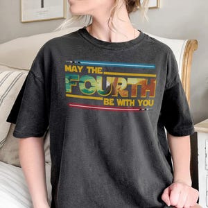May include: Dark grey t-shirt featuring the phrase "MAY THE FOURTH BE WITH YOU" in yellow, blue, and red. The design includes blue and red lightsaber graphics. A casual wear item.