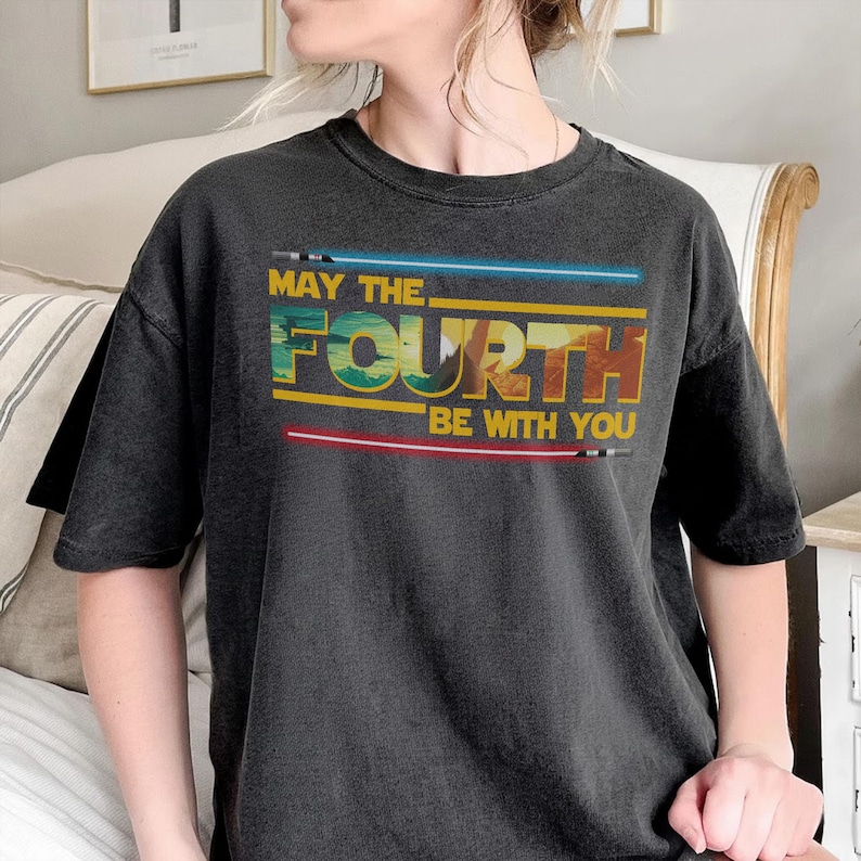 May include: Dark grey t-shirt featuring the phrase "MAY THE FOURTH BE WITH YOU" in yellow, blue, and red. The design includes blue and red lightsaber graphics. A casual wear item.