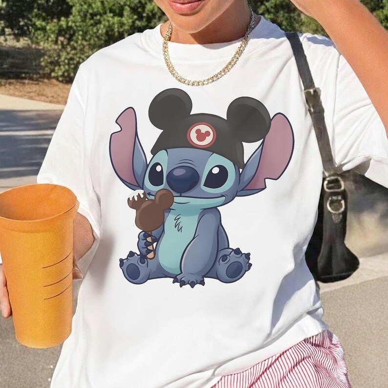 May include: White t-shirt featuring a cartoon illustration of Stitch wearing a Mickey Mouse hat and holding a chocolate ice cream bar. The character is blue with large ears and a friendly expression. The shirt is a casual style.