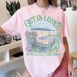 Puede incluir: Camiseta rosa claro con un dise&ntilde;o de carrusel y el texto verde "GET IN LOSER" en la parte superior. Debajo, el texto dice "We're Learning About Lettuce". El carrusel est&aacute; rodeado de flores.
