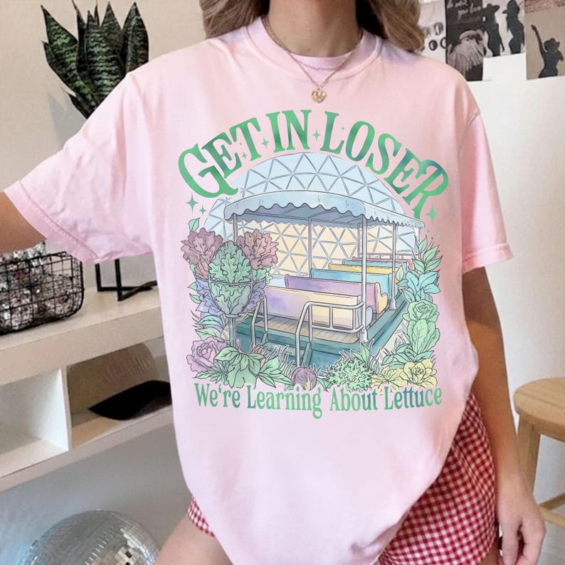 Puede incluir: Camiseta rosa claro con un dise&ntilde;o de carrusel y el texto verde "GET IN LOSER" en la parte superior. Debajo, el texto dice "We're Learning About Lettuce". El carrusel est&aacute; rodeado de flores.