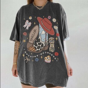 May include: A charcoal gray t-shirt featuring a graphic print of a cowboy boot with a cow print pattern, a red hat, building blocks with the letters T, S, and the number 13, a bow, flowers, and the text "TAYLOR" and "I KNEW IT I KNEW YOU".