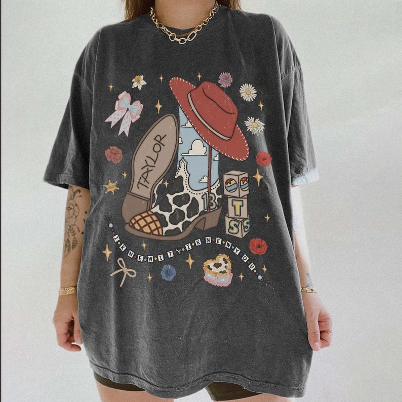 May include: A charcoal gray t-shirt featuring a graphic print of a cowboy boot with a cow print pattern, a red hat, building blocks with the letters T, S, and the number 13, a bow, flowers, and the text "TAYLOR" and "I KNEW IT I KNEW YOU".