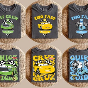 May include: Six dark grey t-shirts with car-themed designs. The shirts feature cartoon cars and text such as "PIT CREW Daddy", "TWO FAST Mommy", and "CHICK HICKS". Each design includes a racing flag and a Route 66 sign.