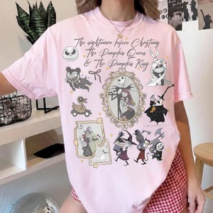 May include: A pale pink t-shirt with illustrations and text from "The Nightmare Before Christmas." The design includes Jack Skellington, Sally, and other characters in a framed portrait and other illustrations. The text reads "The Nightmare Before Christmas, The Pumpkin Queen & The Pumpkin King."