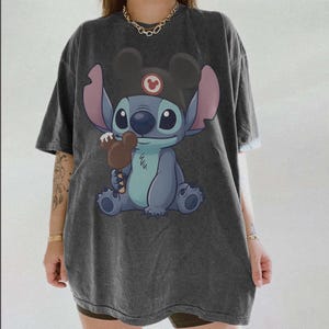 May include: Dark gray t-shirt featuring a cartoon Stitch character wearing a Mickey Mouse hat and holding a chocolate ice cream bar. The graphic is in shades of blue, pink, and brown. The shirt has short sleeves and a relaxed fit.