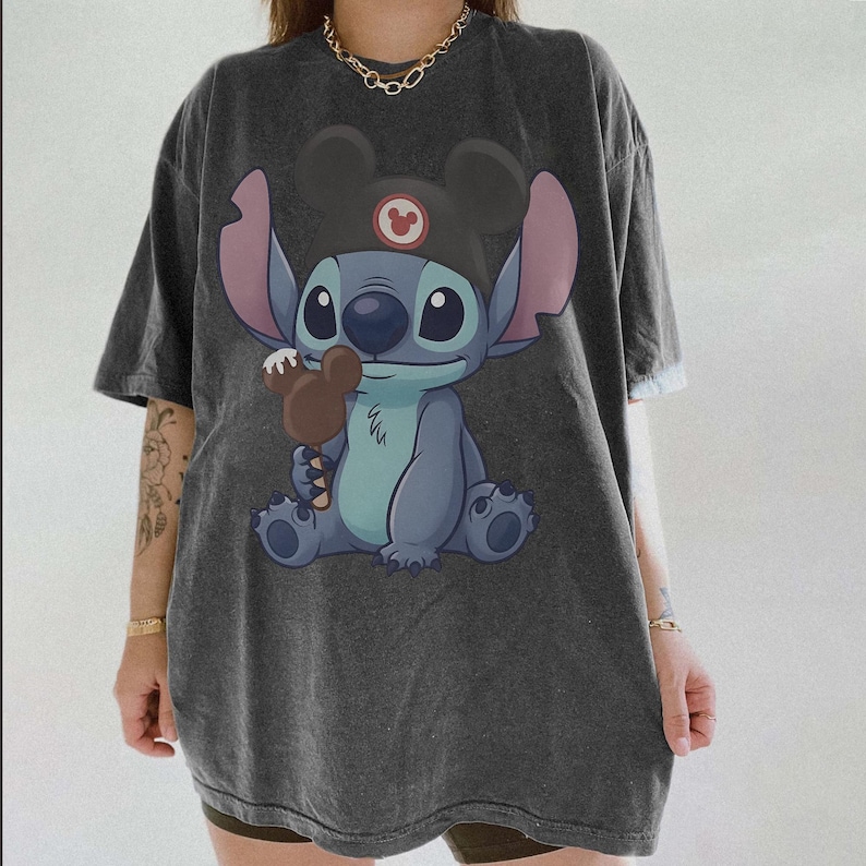 May include: Dark gray t-shirt featuring a cartoon Stitch character wearing a Mickey Mouse hat and holding a chocolate ice cream bar. The graphic is in shades of blue, pink, and brown. The shirt has short sleeves and a relaxed fit.