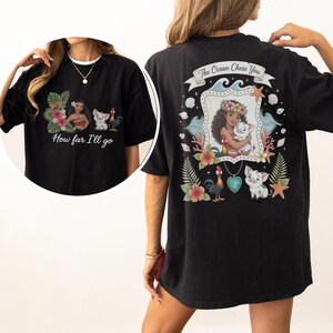 May include: Black t-shirt with a vibrant graphic inspired by an animated film. The front displays a character, a pig, and a rooster with the text "How far I'll go." The back design features the text "The Ocean Chose You."