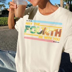 May include: Cream-coloured t-shirt featuring the phrase "MAY THE FOURTH BE WITH YOU" in green, yellow, and brown, with blue and red lightsaber designs. A fun tee for Star Wars fans.