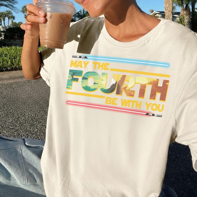 May include: Cream-coloured t-shirt featuring the phrase "MAY THE FOURTH BE WITH YOU" in green, yellow, and brown, with blue and red lightsaber designs. A fun tee for Star Wars fans.