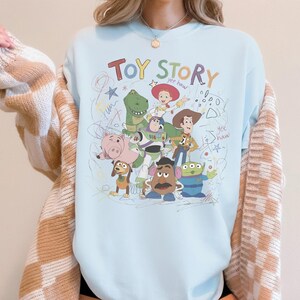May include: A light blue sweatshirt featuring a colorful graphic of Toy Story characters, including Woody, Buzz Lightyear, and Rex. The words "Toy Story" are written in a playful font above the characters, with scribbled accents.