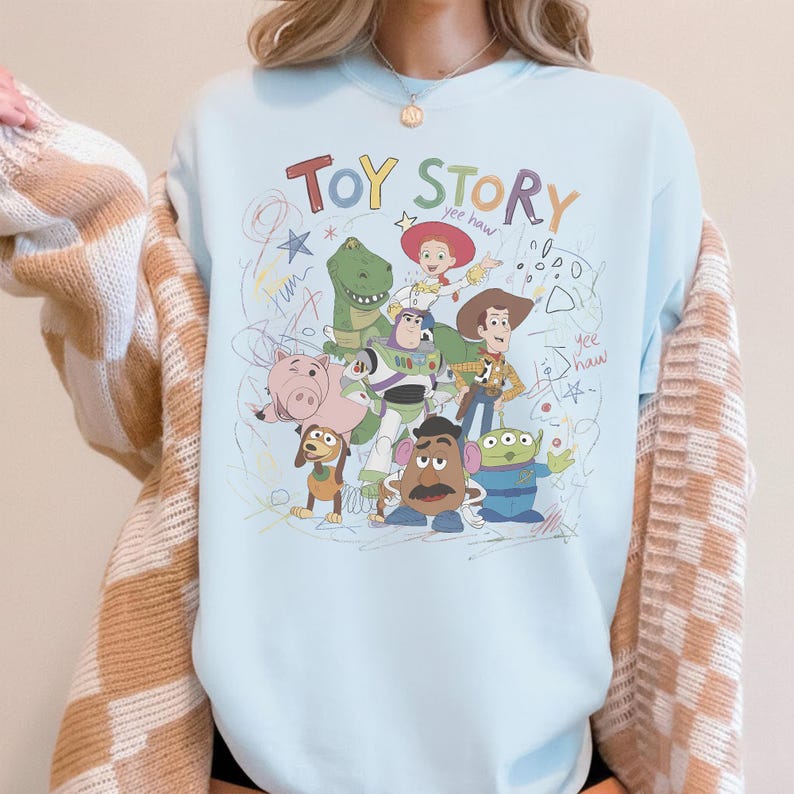 May include: A light blue sweatshirt featuring a colorful graphic of Toy Story characters, including Woody, Buzz Lightyear, and Rex. The words "Toy Story" are written in a playful font above the characters, with scribbled accents.