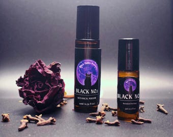 May include: Two glass bottles of "Black No. 1 Botanical Perfume" with black caps, featuring a purple label with a black cat silhouette and moon. A dried burgundy rose and cloves are arranged nearby. Each bottle is 50ml (0.34 fl oz).