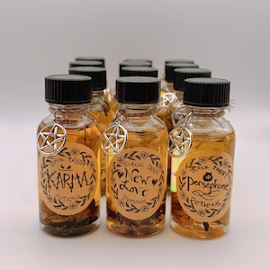 May include: A collection of small clear glass bottles filled with amber liquid and botanical elements. Each bottle has a black screw cap, a silver pentagram charm on a twine loop, and a round tan label reading Black Tabby Potions with names Karma, New Love, or Persephone.