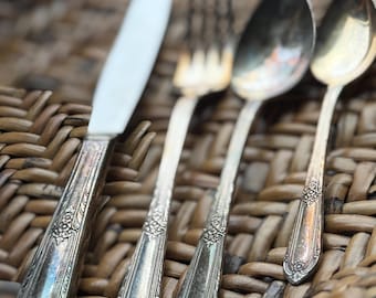 Könnte beinhalten: Ein Satz antiker Silberbestecke, darunter ein Messer, eine Gabel und zwei Löffel, angeordnet auf einer gewebten Oberfläche. Das Besteck hat verzierte florale Details an den Griffen und eine glänzende Oberfläche.