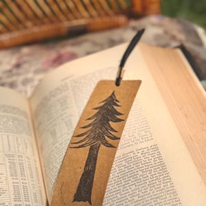 Könnte beinhalten: Handgefertigter Lesezeichen mit einem schwarzen Baumdesign auf braunem Papier. Das Lesezeichen befindet sich in einem offenen Buch. Oben ist eine schwarze Kordel befestigt. Das Buch zeigt gedruckten Text.