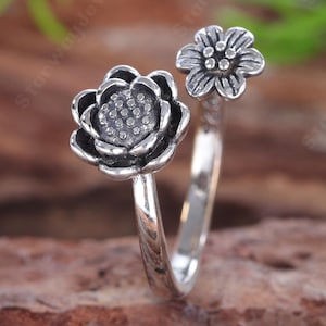 Könnte beinhalten: Ein offener Silberring mit zwei verschiedenen Blumenmotiven. Ein Ende zeigt eine größere Blume mit mehreren Blütenblättern, während das andere Ende eine kleinere Blüte mit sechs Blütenblättern aufweist. Das Metall hat ein oxidiertes Finish.