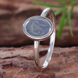 Könnte beinhalten: Ein silberner Ring mit einer ovalen Vorderseite und einer dunklen, vertieften Mitte. In der Mitte des Ovals befindet sich ein eingravierter, kursiver Buchstabe S. Die Ringschiene ist schmal und poliert und weist durchgehend ein metallisches Finish auf.