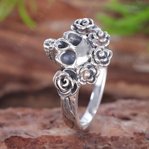 Könnte beinhalten: Ein silberner Ring mit einem zentralen Totenkopfmotiv, das von mehreren skulptierten Rosen umgeben ist. Das Metall hat eine polierte Oberfläche mit abgedunkelten Vertiefungen, um die Details des Totenkopfs und der floralen Elemente hervorzuheben.