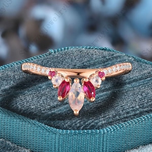 Könnte beinhalten: Ein roségoldener Ring mit einem gebogenen Band, das mit kleinen, klaren Steinen besetzt ist. Der Ring hat einen Cluster aus Edelsteinen, darunter rote, weiße und ein zentraler birnenförmiger Stein. Der Ring wird auf einer tealblauen Samtoberfläche präsentiert.