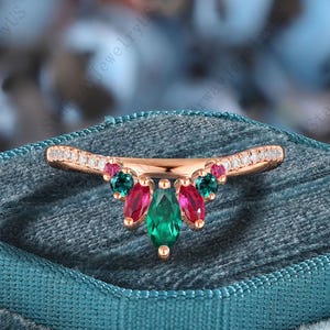 Könnte beinhalten: Ein roségoldener Ring mit einem gebogenen Band, das mit kleinen, klaren Steinen besetzt ist. Der Ring hat einen Cluster aus farbigen Edelsteinen, darunter Grün, Rosa und Türkis, die in einem dekorativen Muster angeordnet sind. Der Ring wird auf einer teal-farbenen Samtoberfläche präsentiert.