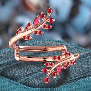 Könnte beinhalten: Ein roségoldener Ring mit Blattdesign, besetzt mit roten Edelsteinen. Der Ring umschlingt den Finger und weist eine astähnliche Struktur mit kleinen, runden und größeren, oval geformten Edelsteinen auf. Der Ring wird auf einer teal-farbenen Samtoberfläche präsentiert.