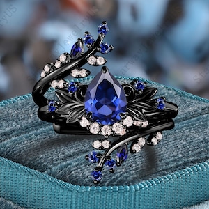 Könnte beinhalten: Schwarzer Ring mit einem tropfenförmigen, tiefblauen Edelstein als Mittelpunkt. Der Ring ist mit kleineren blauen und weißen Edelsteinen und einem floralen Design verziert. Der Ring liegt auf einer teal-farbenen Samtoberfläche.