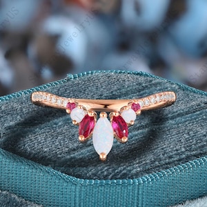 Könnte beinhalten: Ein roségoldener Ring mit einem gebogenen Band, besetzt mit kleinen, klaren Steinen. Der Ring hat eine Ansammlung von Edelsteinen in Weiß, Rot und einen größeren weißen Stein in Marquiseform in der Mitte. Der Ring wird auf einer teal-farbenen Samtoberfläche präsentiert.