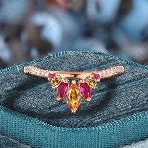 Könnte beinhalten: Ein roségoldener Ring mit einem geschwungenen Band, besetzt mit kleinen, klaren Steinen. Der Ring hat einen Cluster aus marquiseförmigen Edelsteinen in Rot-, Gelb- und Orangetönen. Der Ring wird auf einer teal-farbenen Samtoberfläche präsentiert.