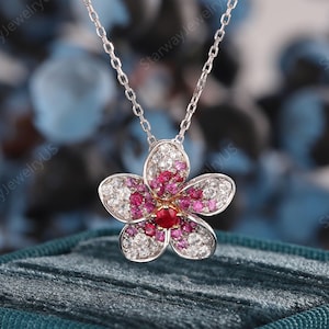 Könnte beinhalten: Eine silberne Blumenanhänger-Halskette mit Kette. Der Anhänger hat ein Blumenmuster mit einem roten Mittelstein, umgeben von rosa und weißen Edelsteinen. Die Halskette wird auf einer teal-farbenen Samtoberfläche präsentiert.