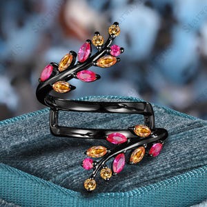 Könnte beinhalten: Ein schwarzer Ring mit Blattmuster, verziert mit rosa und gelben Edelsteinen. Der Ring liegt auf einer tealblauen Samtoberfläche, die seine detaillierte Gestaltung und leuchtenden Farben hervorhebt. Ein einzigartiges und elegantes Schmuckstück.