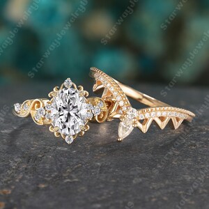 Könnte beinhalten: Ein goldfarbenes Verlobungsring-Set. Der Verlobungsring hat einen Diamanten im Marquise-Schliff als Mittelstein, umgeben von kleineren Diamanten. Der Ehering hat ein geschwungenes Design mit kleinen Diamanten. Die Ringe liegen auf einer dunkelgrauen Oberfläche.