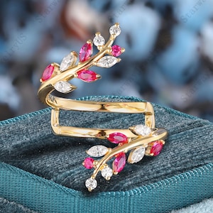 May include: A gold-colored ring with a unique design, featuring ruby and diamond accents. The ring has a wrap-around style with leaf-like patterns adorned with red rubies and white diamonds. The ring is displayed on a teal velvet surface.