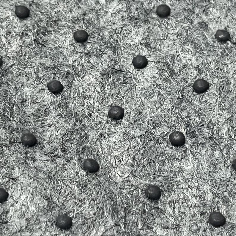 May include: A textured, fibrous gray surface featuring a repeating pattern of small, dark, circular studs arranged in a grid. The material appears to have a coarse, woven, or matted consistency with a monochromatic, high-contrast appearance.
