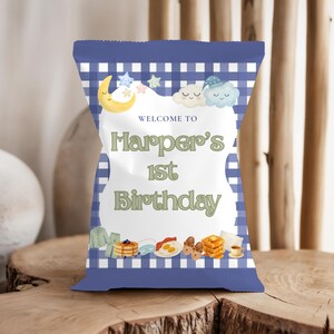 May include: A blue and white gingham-patterned party favor bag with the text "Welcome to Harper's 1st Birthday." The bag features illustrations of a crescent moon, stars, clouds, and breakfast foods.
