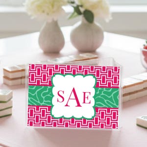 Puede incluir: Una caja de acrílico transparente con un panel frontal decorativo. El panel presenta un patrón geométrico rosa y blanco, una franja verde estampada y un marco festoneado blanco con las letras "SAE" en rosa. La caja está sobre una superficie rosa.